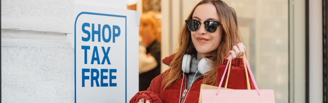 Woman wearing sunglasses and carrying shopping bags by sign that reads “Shop Tax Free”