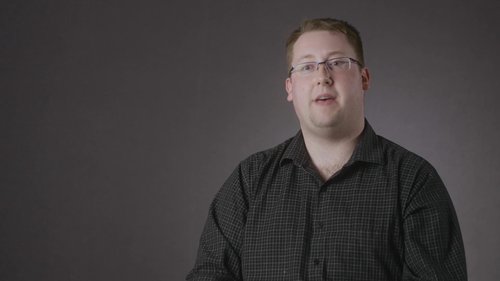 A person wearing a dark checkered shirt against a plain gray background.