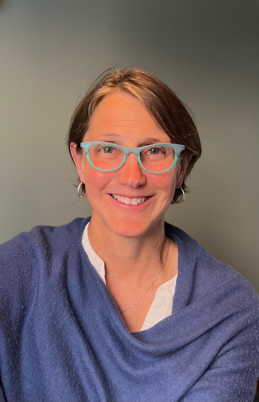 Portrait headshot of a smiling person with short brown hair, blue glasses, hoop earrings, and a blue shawl, set against a plain background.