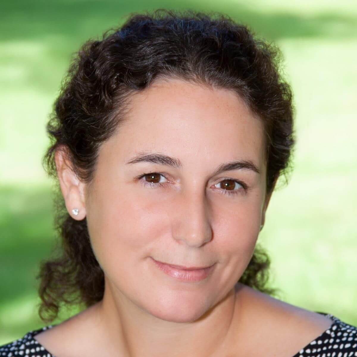 A close-up, professional headshot of a woman with curly brown hair, wearing a black and white top, photographed outdoors with a green blurred background.