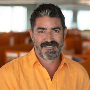A person wearing an orange shirt in an office setting with cubicles and bright windows in the background.