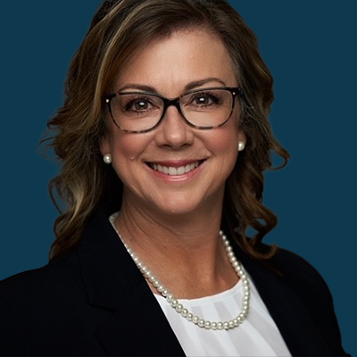 Professional studio headshot of Christine Martin against a dark teal background. She wears glasses, a dark blazer, white blouse, and pearl jewelry, conveying a confident and approachable corporate presence. Suitable for webinar speaker profiles and bio sections.